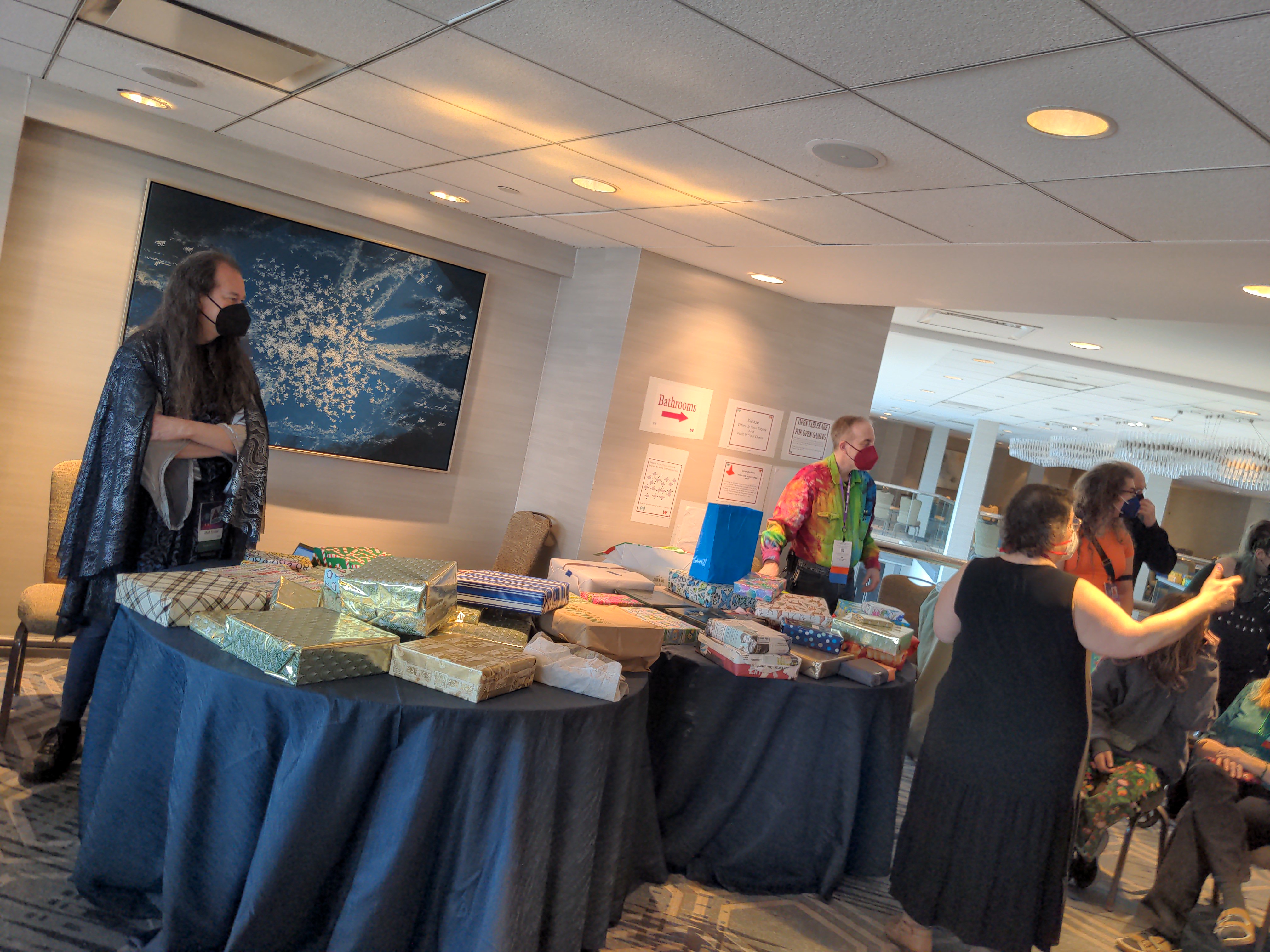 A table stacked high with wrapped board games. Several people in covid masks and dressed in geeky finery stand around coordinating the game.
